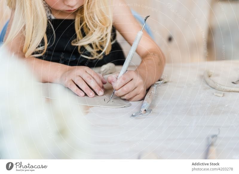 Unrecognizable kid cutting clay piece in studio girl pottery loop hobby workshop instrument child craft black apron table sharp light handmade creative skill