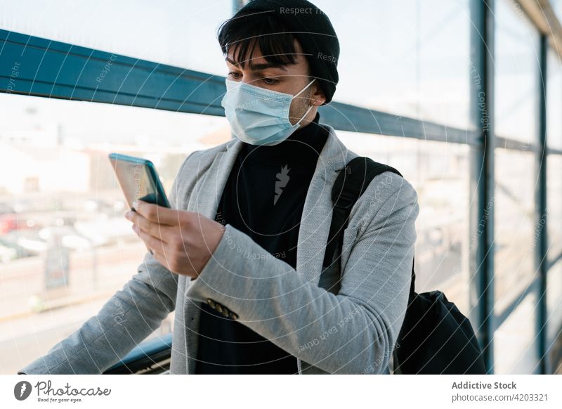 Traveling man with backpack using smartphone at railway station student browsing corridor hallway male walk mask protect commute prevent safety gadget device