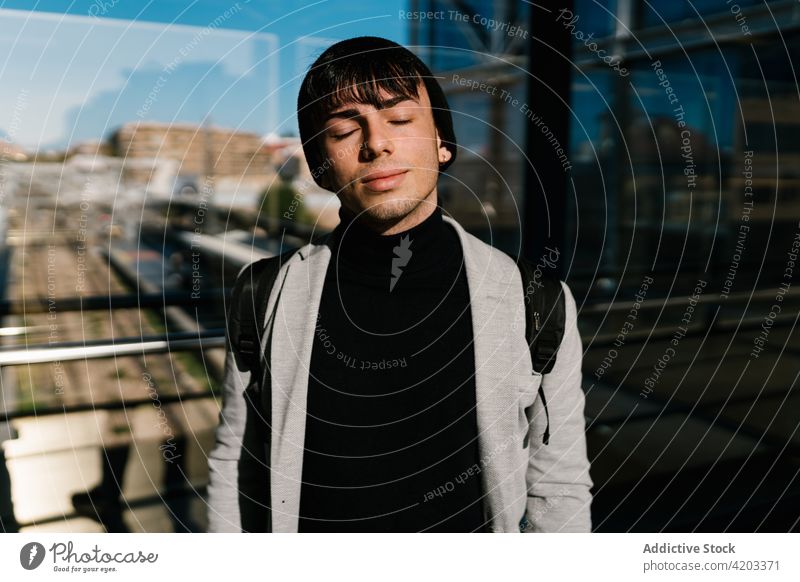 Stylish man standing near glass wall lit by sunlight serene style harmony student tranquil male backpack young city handsome calm street hipster dreamy daydream