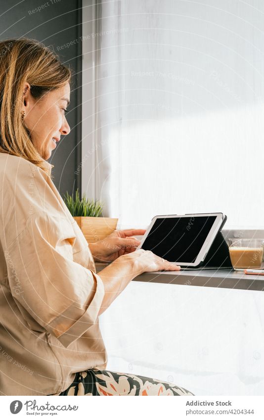 Smiling woman using tablet at counter in kitchen home mature browsing smile device female morning gadget middle age social media connection surfing internet