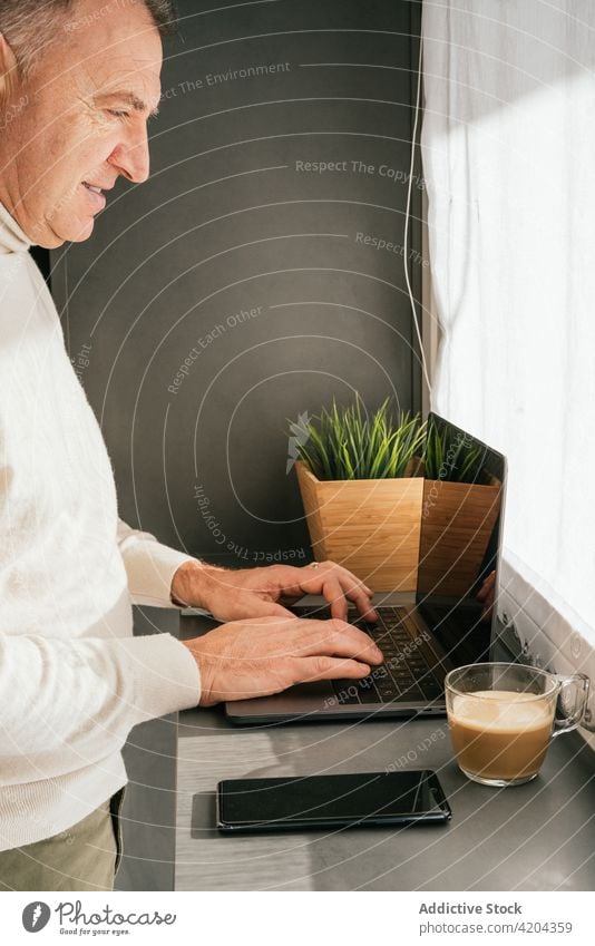 Cheerful mature man siting on chair at counter with laptop in kitchen morning smile home breakfast coffee male work cup netbook remote cheerful happy middle age