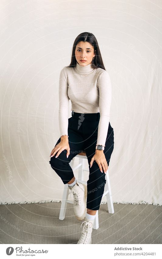 Young woman sitting on stool in light studio background solitude touch head depression room casual cloth thoughtful pensive female alone sensitive reflective