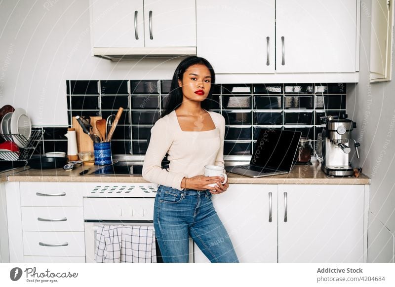 Confident young Hispanic woman standing with cup in modern kitchen laptop confident drink contemporary beverage at home domestic female hispanic counter