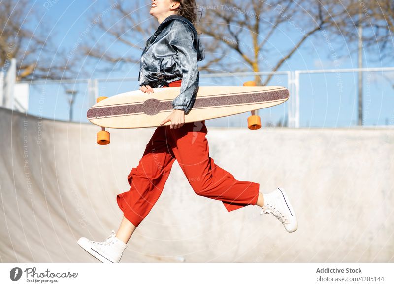 Excited woman with longboard jumping against leafless trees in town excited happy energy stylish fence blue sky cloudy athlete trendy style cheerful smile enjoy