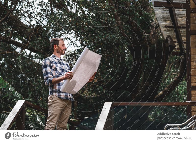 Male architect with blueprint standing near modern house man building project draft paper terrace wooden male cottage construction forest guy estate