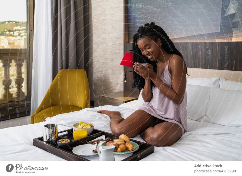 Happy young black lady having breakfast in cozy hotel room woman drink coffee bed content smile comfort tourist relax delicious female african american ethnic