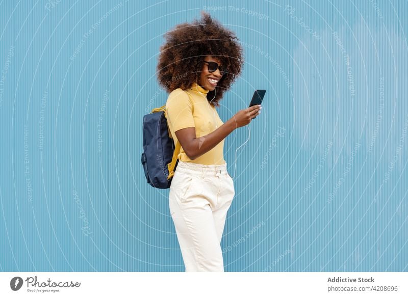 Cheerful black woman browsing smartphone near blue wall selfie moment content headphones portrait using gadget colorful device self portrait happy spare time