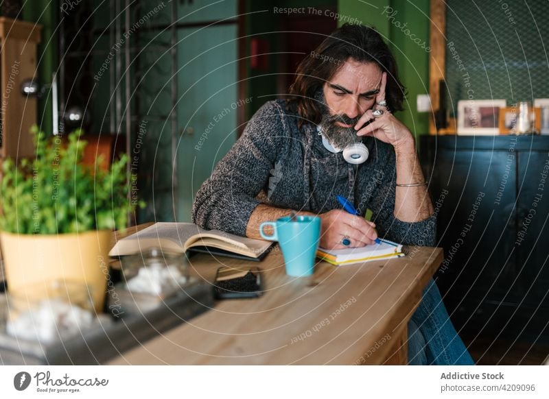 Pensive Hispanic writer taking notes on paper at home take note thoughtful touch temple work professional man book house literature knowledge hot drink tea