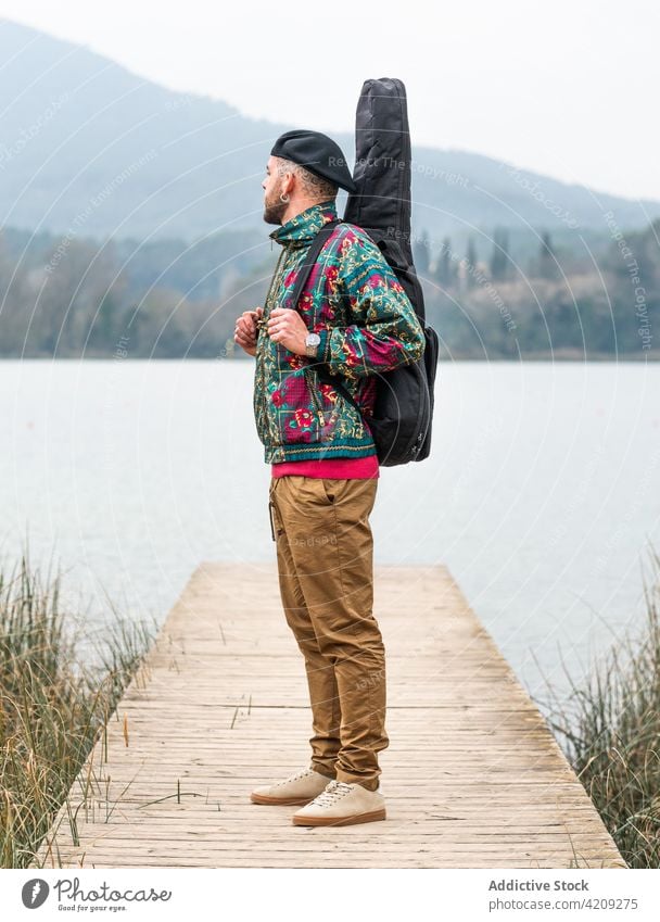 Pensive man standing with guitar against calm lake and mountain guitarist musician nature enjoy pensive thoughtful male wooden pier river cloudy water tranquil