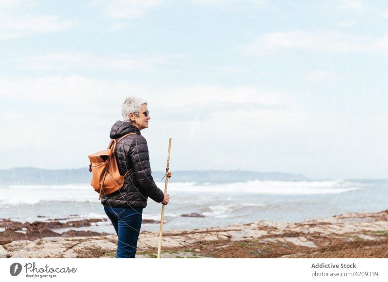 Senior trekker walking on rocky sea beach during trip boulder pole explore wanderlust woman seascape nature sunglasses backpack hike uneven coast ocean stormy