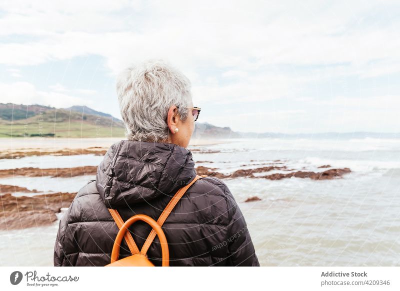 Anonymous senior hiker admiring sea under cloudy sky admire ocean trek enjoy nature seascape woman contemplate pole trekker trip explore wanderlust sunglasses