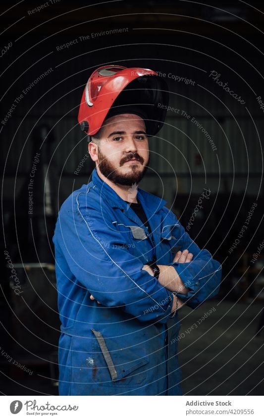 Confident male mechanic in helmet and overall in workshop man workman serious hardhat garage worker professional adult job blue uniform repairman stand beard