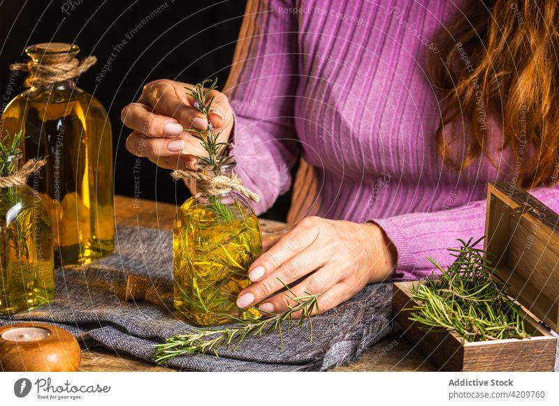 Unrecognizable female demonstrating bottle with oil and rosemary woman herb demonstrate cloth table glass show delicate essential light purple casual creative