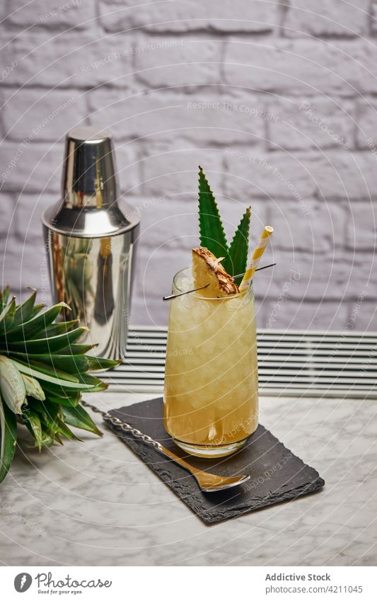 Glass with pineapple cocktail near shaker ice slate cube straw counter spoon leaf glassware creative fresh paper coaster exotic ingredient cold fruit yummy