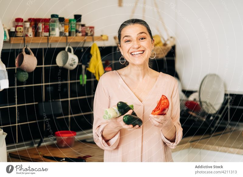 Happy mature woman with vegetables in kitchen bell pepper cucumber leek dishware food ingredient counter healthy food middle age happy smile cheerful positive