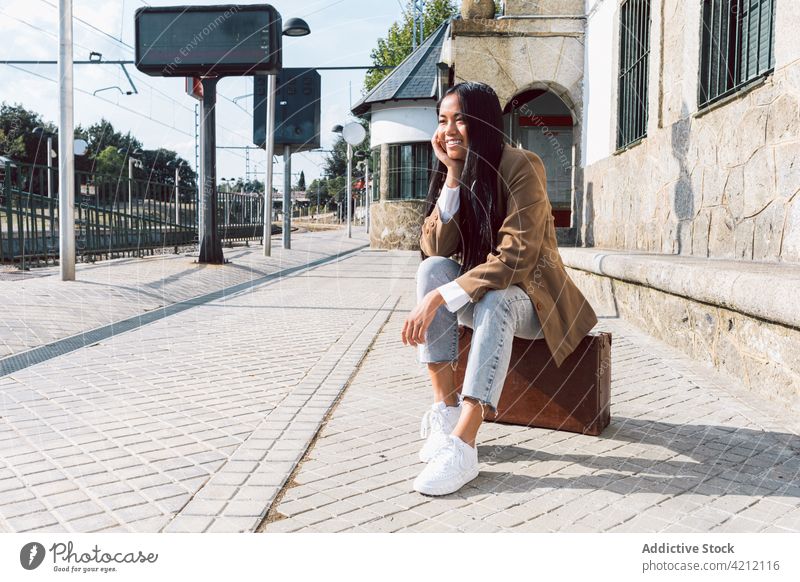 Content Asian woman sitting on vintage suitcase at railway station wait retro traveler platform cheerful female asian ethnic trip departure happy passenger