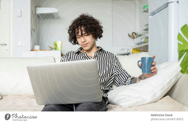 Tattooed man surfing internet on laptop on sofa at home online free time tattoo using gadget device watching coffee cup browsing rest beverage netbook couch