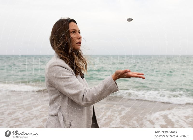 Serene woman tossing stone at seaside seashore autumn beach serene season female cloudy weather calm water nature tranquil harmony fall outerwear coast overcast