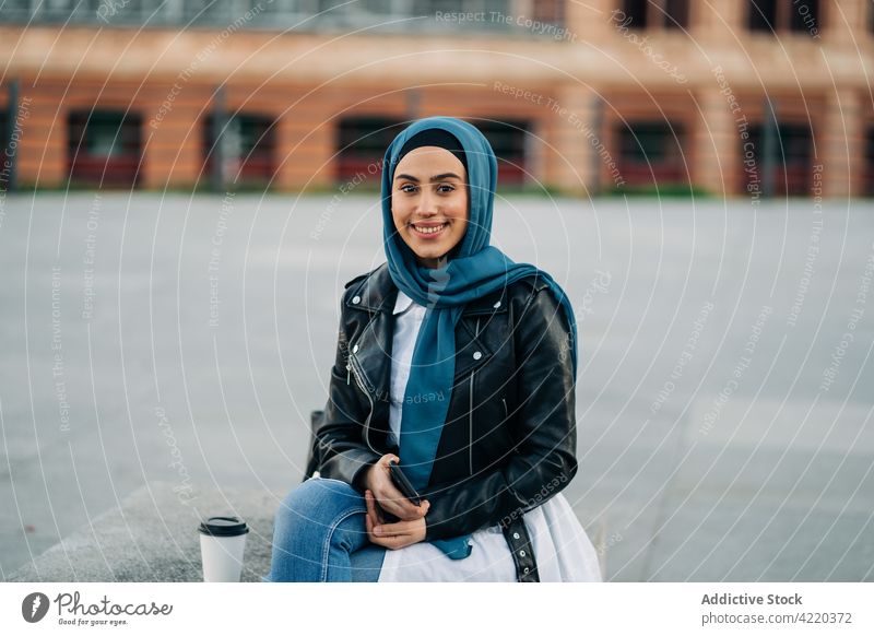 Stylish ethnic woman in hijab holding smartphone on street using surfing city style bench female muslim takeaway drink to go coffee gadget device beverage