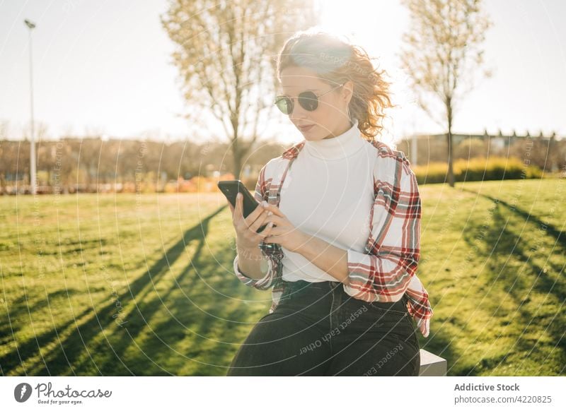 Stylish woman browsing on park smartphone female gadget device mobile using modern style trendy curly hair watching sit bench sunglasses text messaging blonde