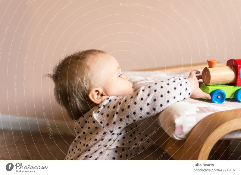 Toddler girl reaching up to grab a colorful wooden train toy; peach and white nursery child toddler fine motor skills pull 10 months old baby young pull up