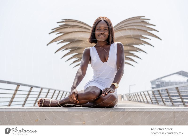 Gorgeous black woman sitting on promenade near wing shaped sculpture gorgeous style elegant content embankment white dress feminine sunny daylight short hair