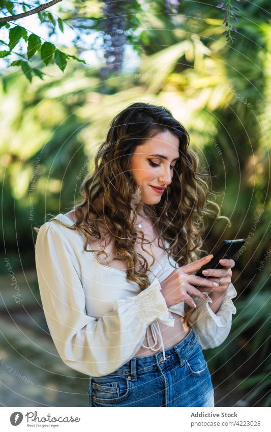 Smiling young woman using smartphone in lush park browsing toothy smile garden content style carefree mobile attractive beautiful cheerful gadget positive