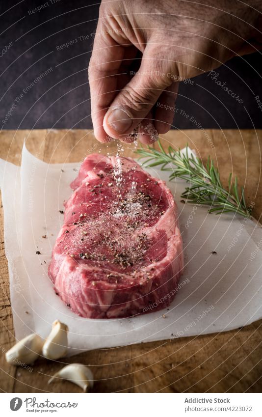 Crop chef salting raw beef steak on parchment seasoning meat protein culinary recipe man process spice rosemary sprig fresh herb clove garlic vegetable prepare