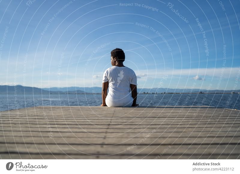 Unrecognizable black woman sitting on pier near rippling lake nature pond contemplate waterfront scenic observe quiet enjoy admire pure picturesque aqua quay