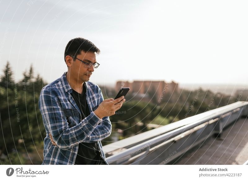 Focused man using smartphone on rooftop browsing suburb focus gadget concentrate message casual modern surfing online social media sunny serious thoughtful male