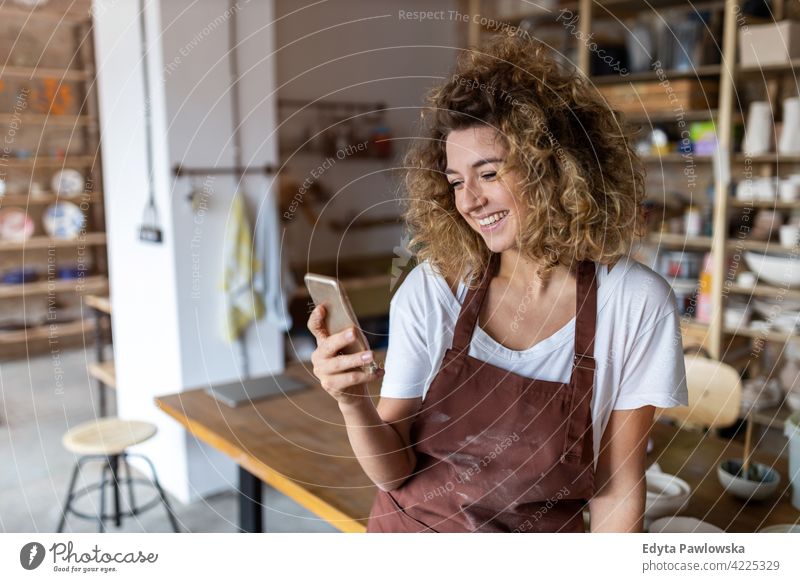 Female potter with smartphone in art studio pottery artist ceramics work working people woman young adult casual attractive female happy Caucasian enjoying