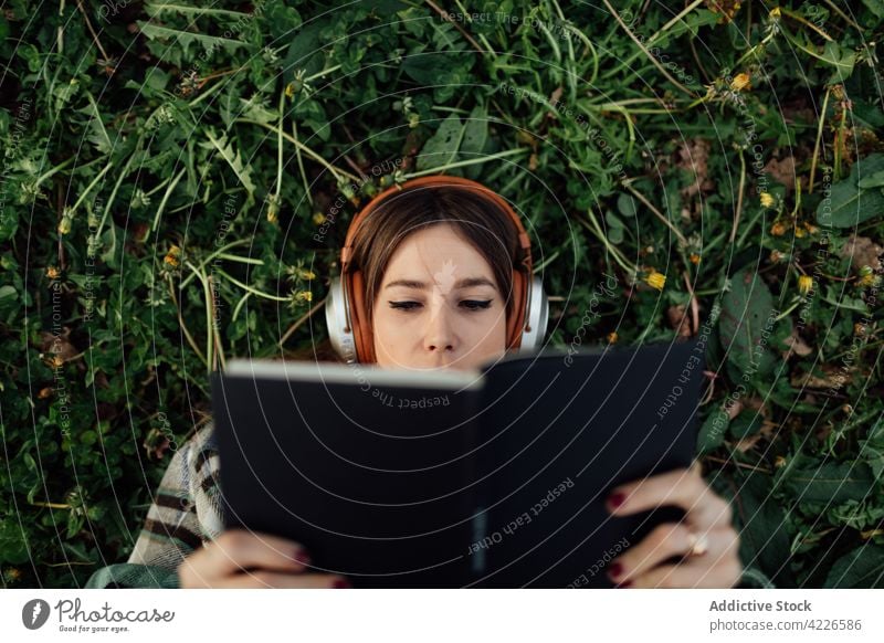 Woman in headphones reading book on grass woman literature knowledge spare time listen using gadget device textbook education music melody millennial attentive