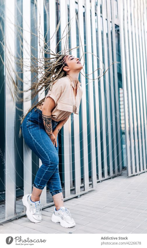 Trendy woman with flying hair on city street fashion style individuality cool tattoo braid enjoy town lean forward wall sneakers pavement pleasure tiptoe denim