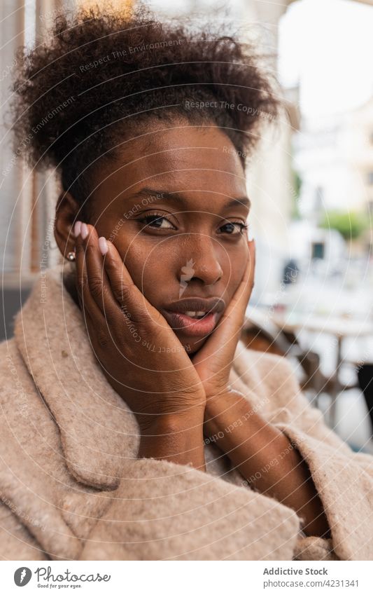 Young black woman holding face in hands on street touch cheek coquette coat positive outerwear appearance young touch face curly hair city casual glad lifestyle