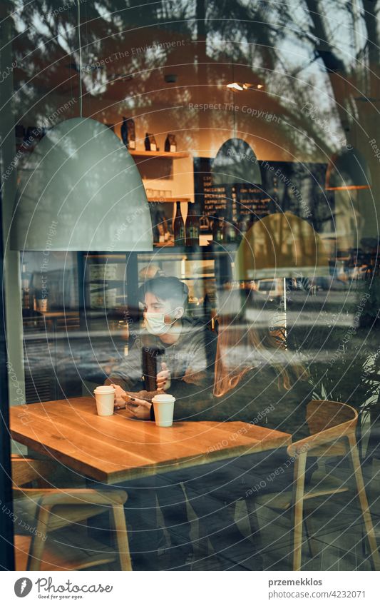 Couple relaxing in a cafe, using smartphone, having a chat, talking together, drinking coffee. Young man and woman having a break, spending time together wearing face masks