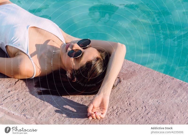 Sensual tourist in swimsuit resting against swimming pool woman lying fashion style sensual sunglasses feminine body vacation travel swimwear water ripple shiny