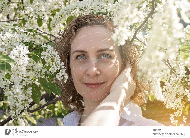 Beautiful smiling caucasian woman with curly hair is posing under a blooming tree. Springtime photoshoot, sunny day. Portrait of a happy woman. Spring sunshine