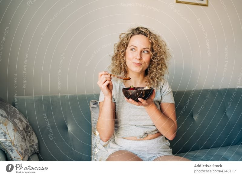 Happy woman eating oatmeal on cozy coach cheerful sofa bowl toothy smile happy carefree comfort delicious satisfied legs crossed friendly couch content food