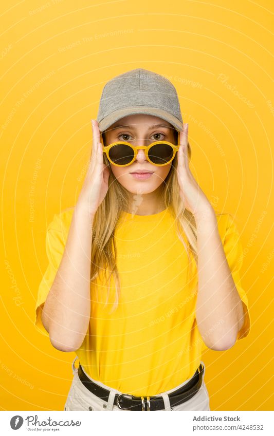 Woman with sunglasses in hands standing against yellow wall woman style vivid hipster street style cool appearance accessory sweater cap young funny bright