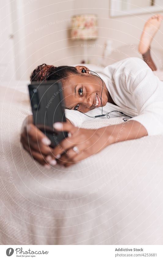 Positive young black woman taking selfie and listening to music while lying on bed smartphone self assured smile relax enjoy recreation content female ethnic