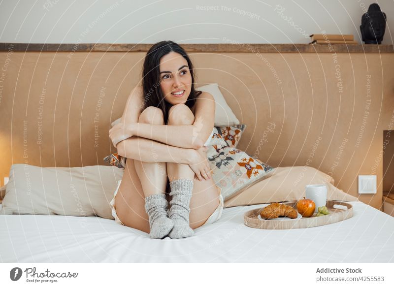 Smiling woman embracing knees and sitting on bed dreamy smile nightwear bedroom morning comfort serene lifestyle attractive brunette peaceful tender gentle