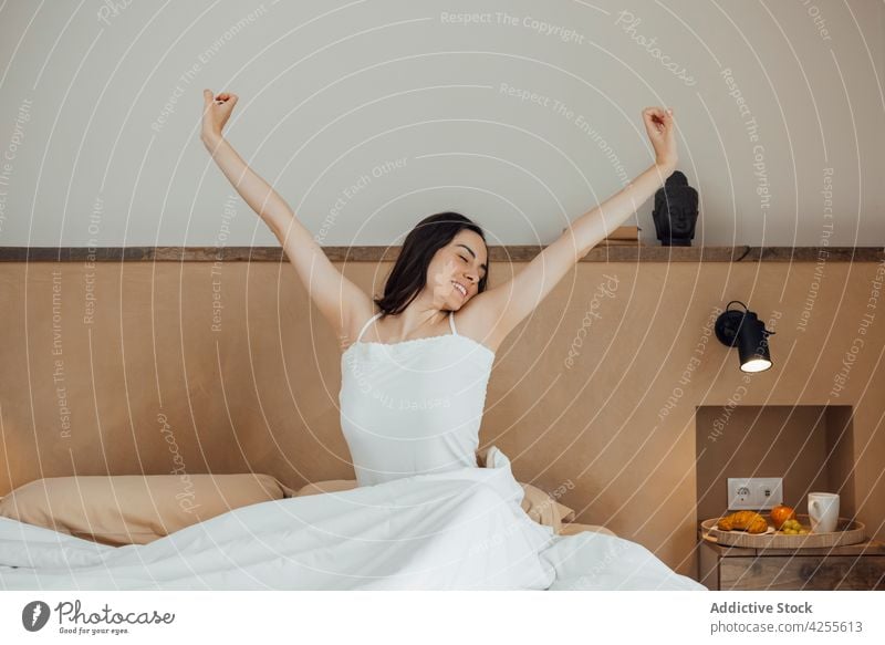 Smiling woman stretching arms after good sleep on bed wake up eyes closed toothy smile content awake bedroom morning glad comfort rest lifestyle cozy young