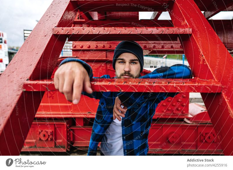 Man in trendy checkered blue shirt leaning on metal construction man checkered shirt confident casual outfit cool rest urban male hat contemporary weekend young