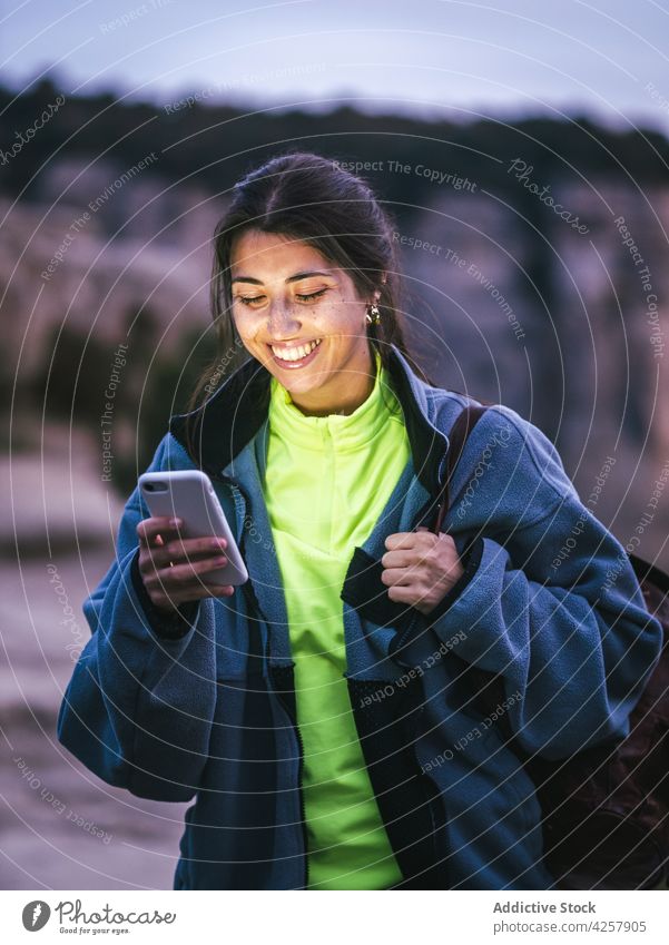 Cheerful young ethnic woman smiling and using smartphone on rocky cliff message smile canyon traveler trekking mountain enjoy rest female hispanic positive