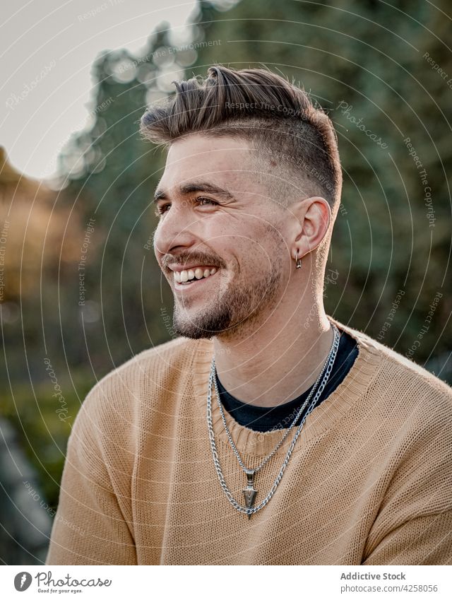 Smiling young man sitting in lush park toothy smile happy casual cheerful appearance glad nature garden beard handsome good looking personality content sweater