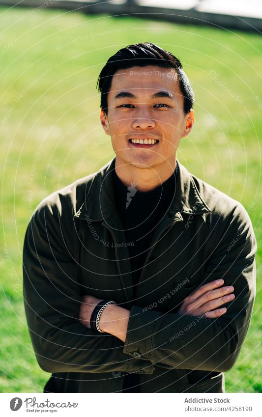 Trendy young ethnic guy standing on lawn in park on sunny day man style self assured smile happy confident delight satisfied free time arms folded fashion alone