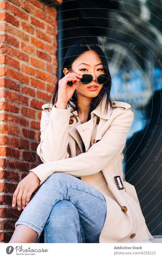 Stylish young ethnic female millennial adjusting sunglasses on street woman style self assured confident trendy rest city serious calm asian brunette coat