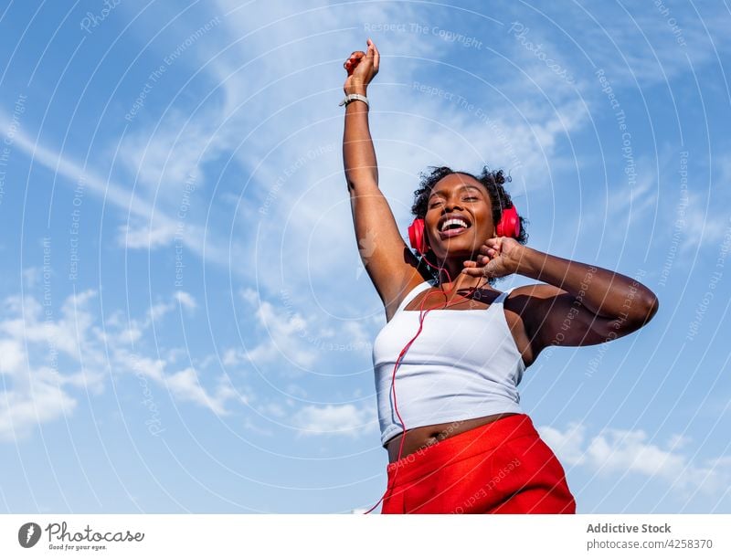 Excited black woman listening to music in headphones and dancing dance joy having fun blue sky melody enjoy arms raised happy young fit energy positive