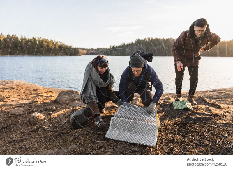 Happy campers putting folding mat on lake coast people trekking friend shore adventure travel nature traveler autumn journey trip millennial warm clothes