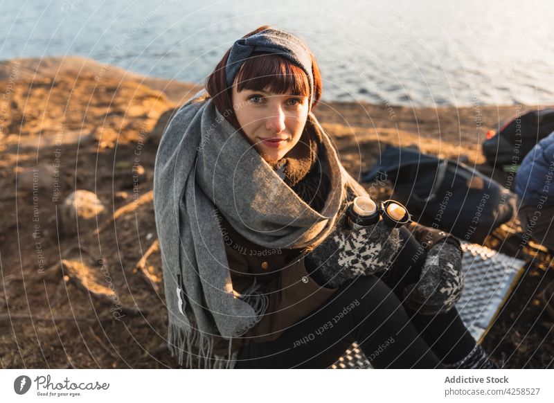 Positive young female hiker drinking hot beverage at campsite near lake woman hot drink camper content smile nature enjoy travel rest warm clothes relax trip